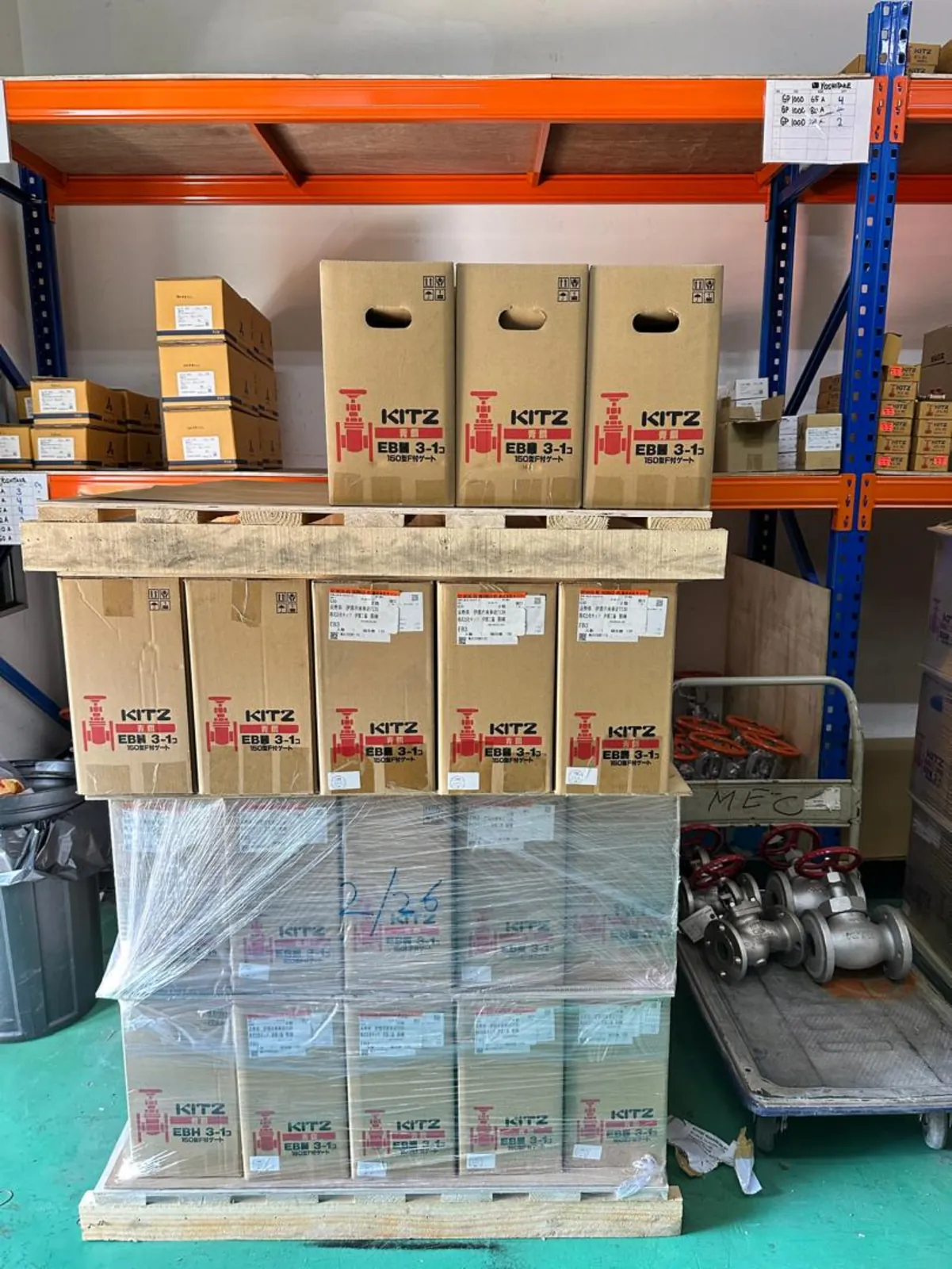 Palletised cartons of flanged gate valves stacked in front of warehouse racking, loose flanged stock on a trolley behind
